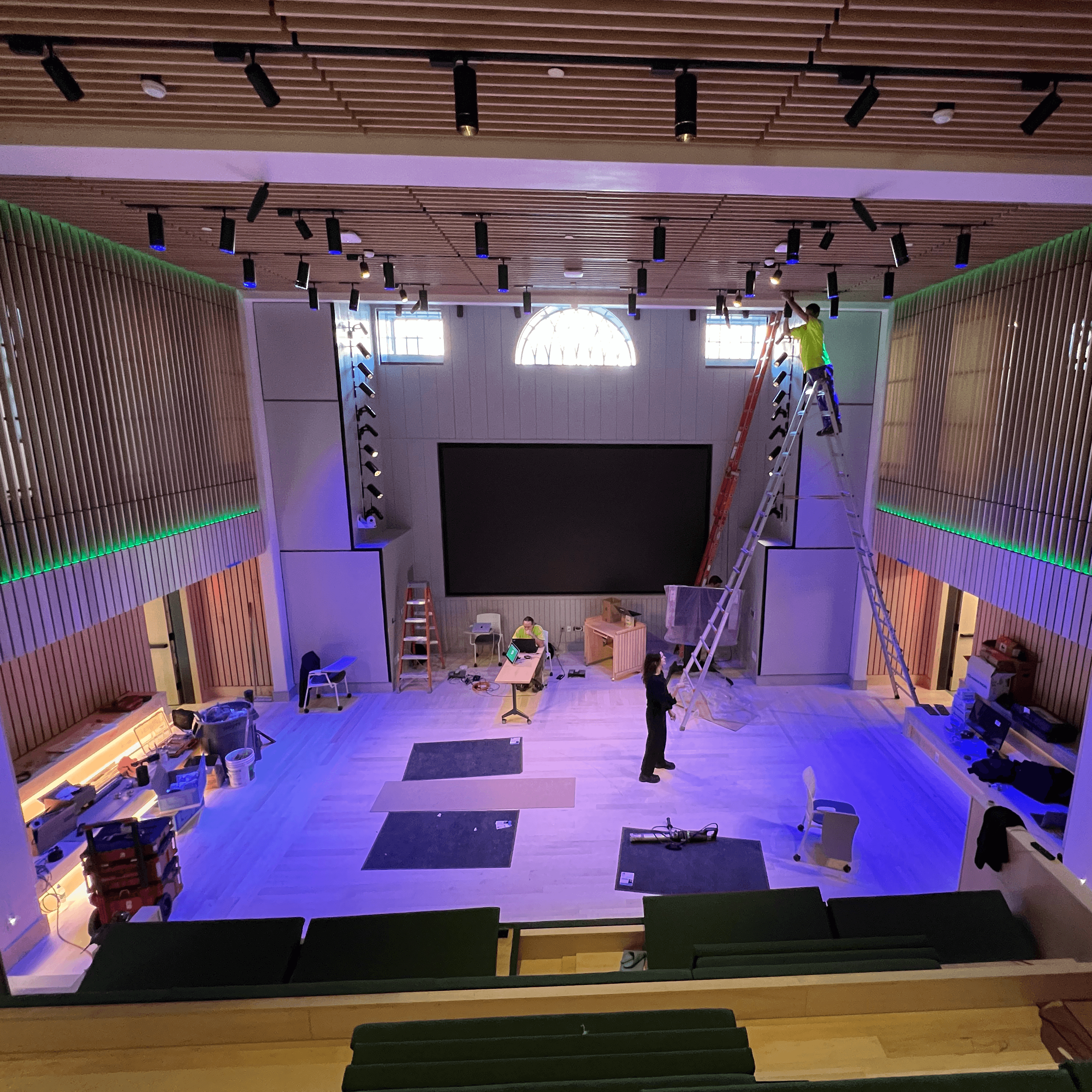 Auditorium under construction with workers on ladders adjusting ceiling-mounted lighting, a large black screen on the far wall, and tools and materials scattered across the floor; linear LED accent lights illuminate the wood slat walls with a green glow.