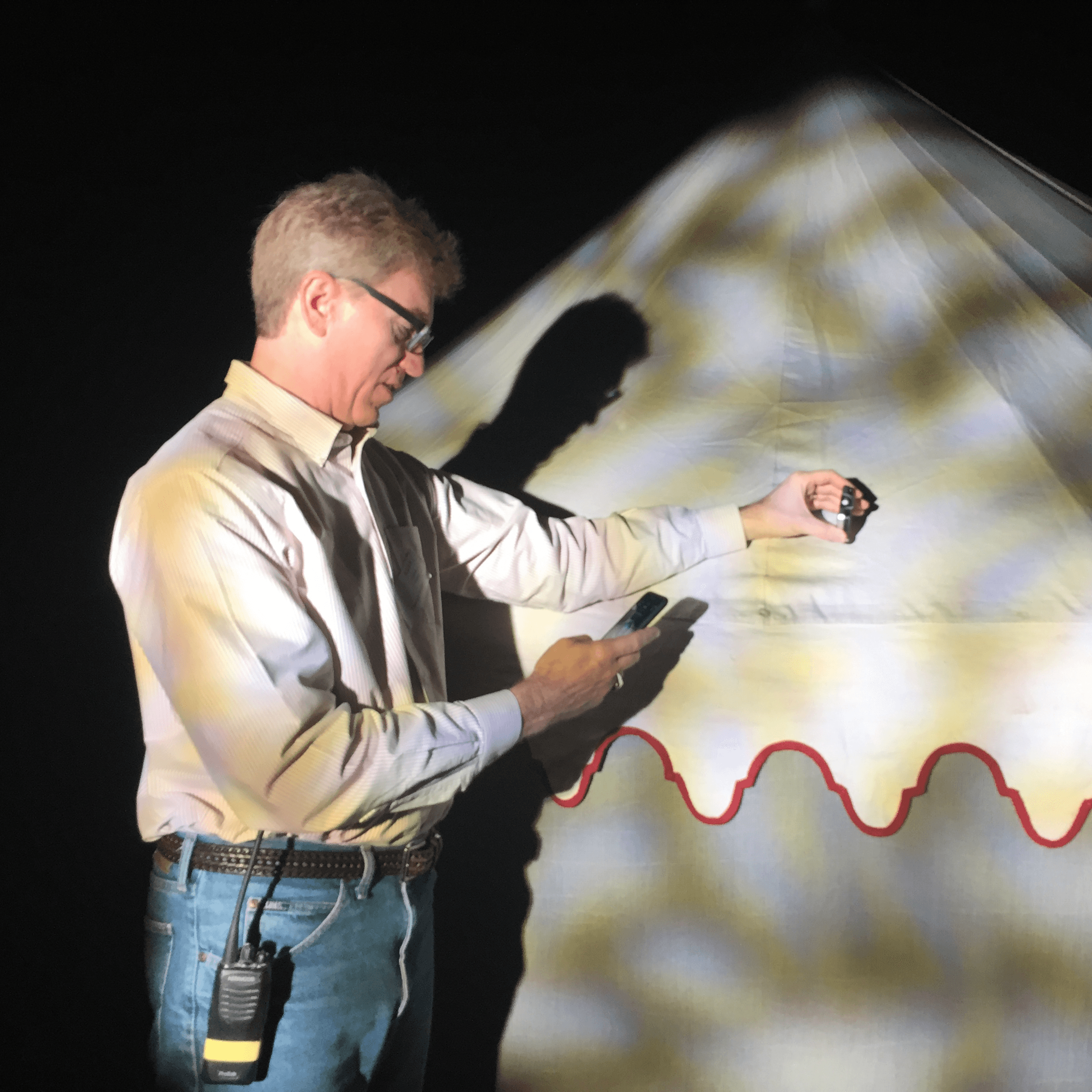 Available Light - Museum - Onsite Commissioning A man with a radio clipped to his jeans uses a handheld device to measure light levels on a fabric surface, which is illuminated by patterned theatrical lighting in a dark environment.