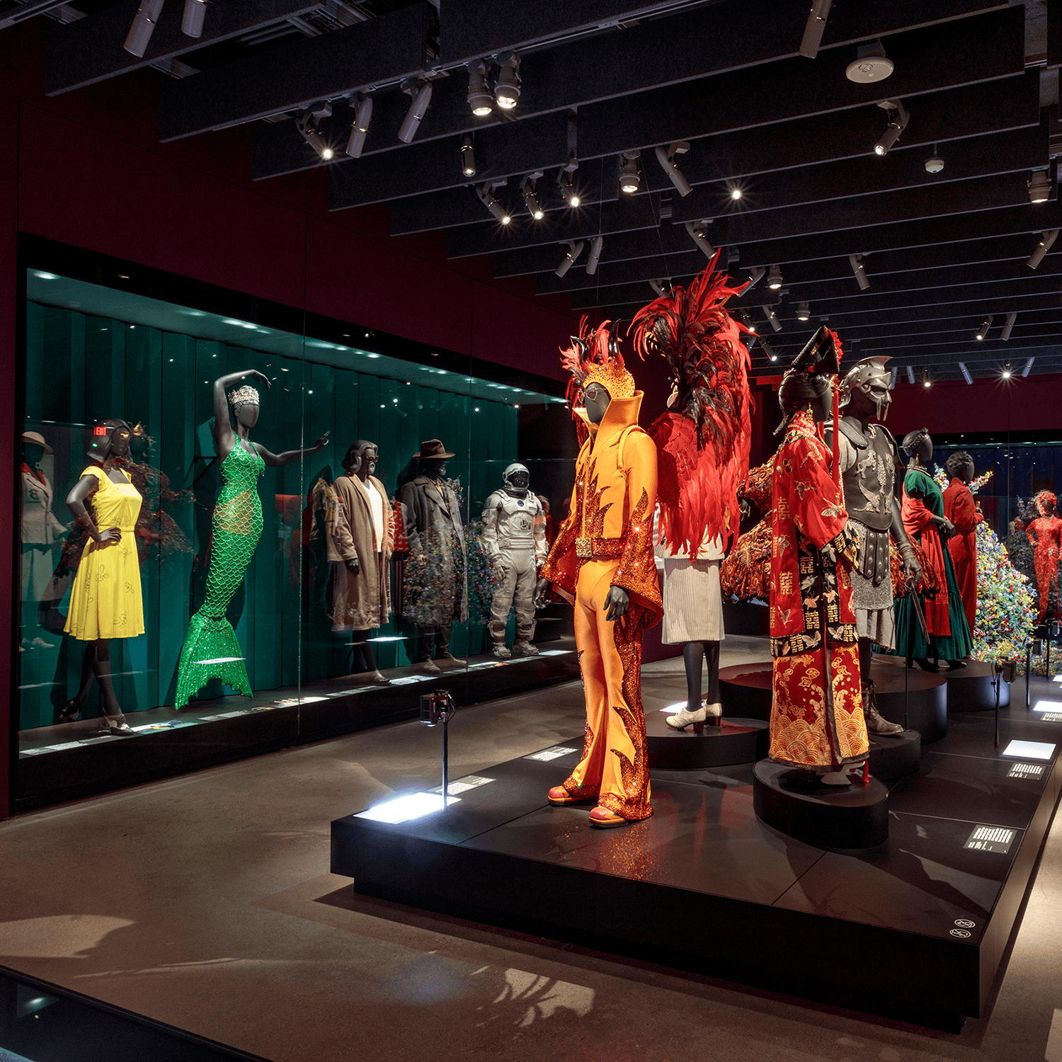 Available Light Museum Post Opening Museum exhibit of theatrical and film costumes displayed on mannequins under dramatic spotlights, featuring colorful and ornate outfits in a dark gallery with glass cases and overhead track lighting.