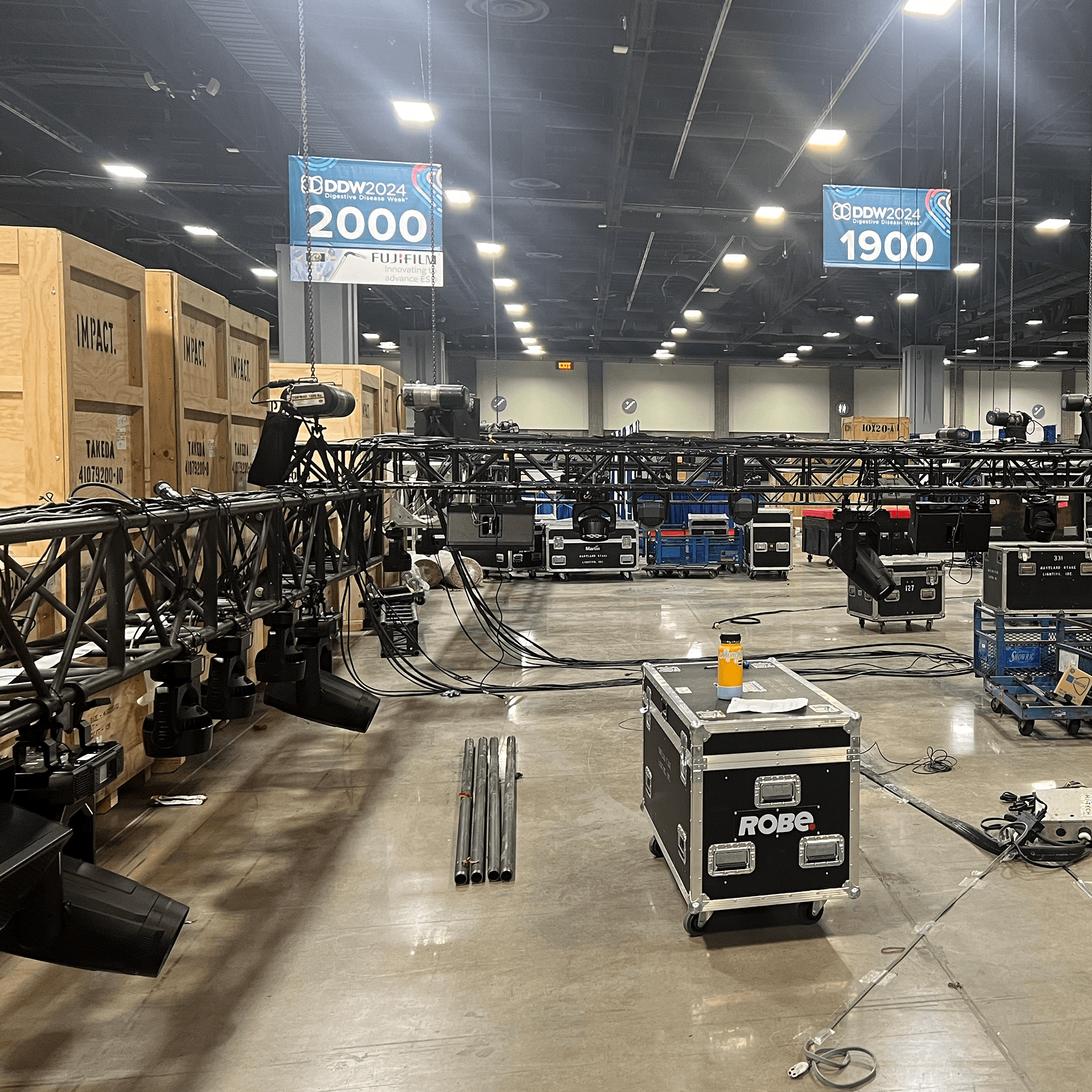 Available Light - Trade Shows - Logistics & Equipment Trade show setup in progress for DDW 2024, showing suspended lighting trusses, crates, equipment cases, and hanging booth signs for exhibitors Fujifilm and others in a large convention hall.