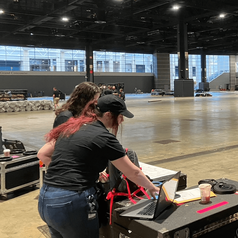 Available Light - Trade Shows - Onsite Support Two team members in black shirts review plans on a laptop inside a large convention hall during trade show setup, with gear cases, coffee cups, and scattered equipment visible in the background.